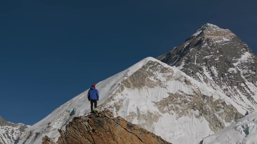 A person stands on a summit and looks at huge mountains