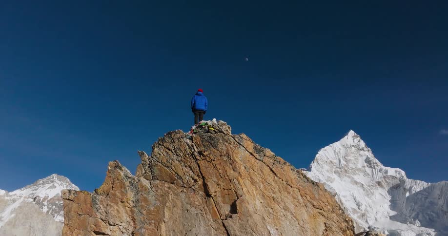A lone individual stands atop a rocky peak surrounded taller mountains