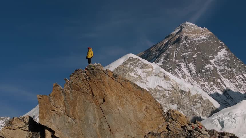 A person in a yellow jacket climbing a large rock and behind him a huge mountain covered in snow