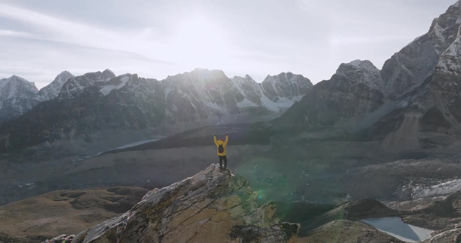Hiker wearing a red hat a yellow coat and a black backpack lifts his hands up as he looks at distant high mountain range