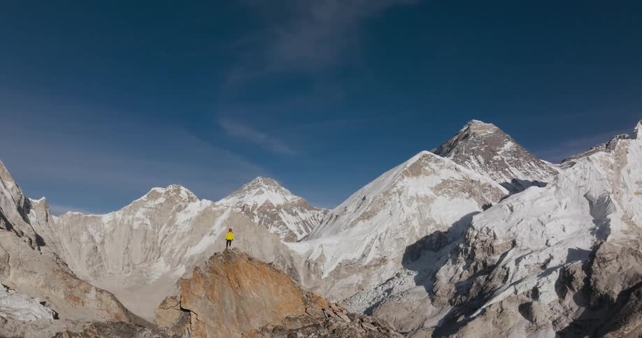 A person in a yellow jacket on top of a mountain and behind there are bigger mountains with snow on top