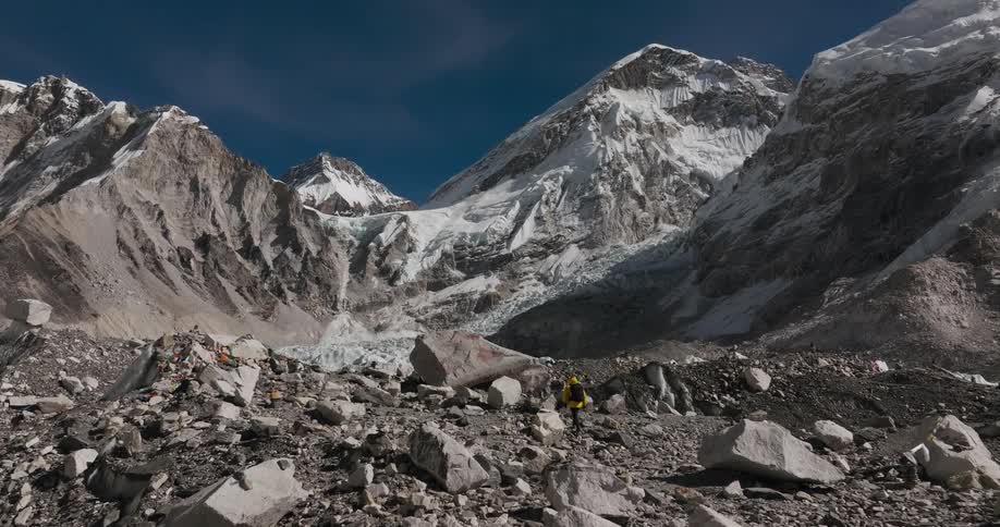 Aerial of man wearing a red hat a yellow coat and a black backpack walks in rough terrain with mount everest in the background