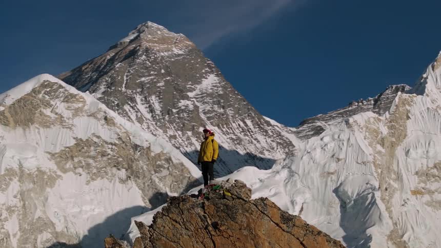 A person in a yellow jacket is standing on top of a rock looking around at the snowcovered mountains