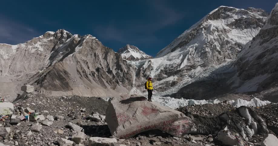 Person climbing on top of a rock overlooking a landscape of snowcapped mountains on a dark blue sky day