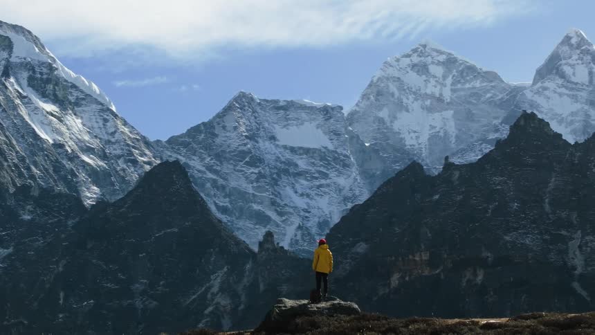 An aerial view captures a man wearing a red hat and a yellow coat standing on a rock gazing at mount everest