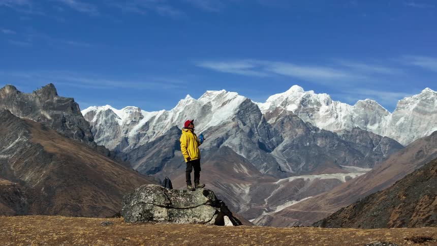 A man stands on a rock and drinks from a bottle against a backdrop of a mountain range