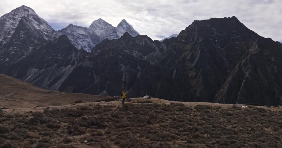 Aerial of man wearing a red hat a yellow coat and a black backpack walks along a ridge with mount everest in the background