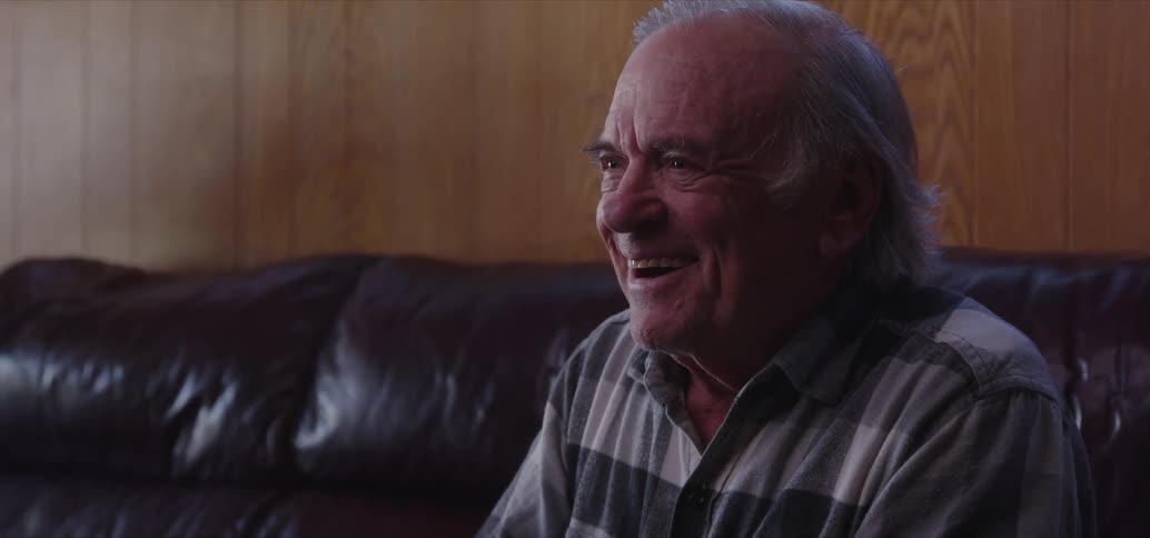 An old man sits on a couch and speaks and laughs