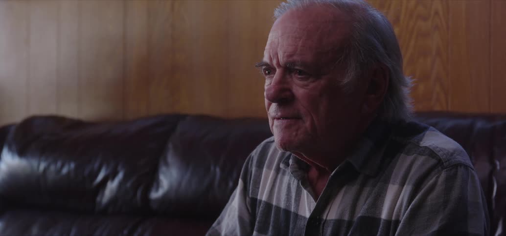 An old man sits on a couch listening to someone speak