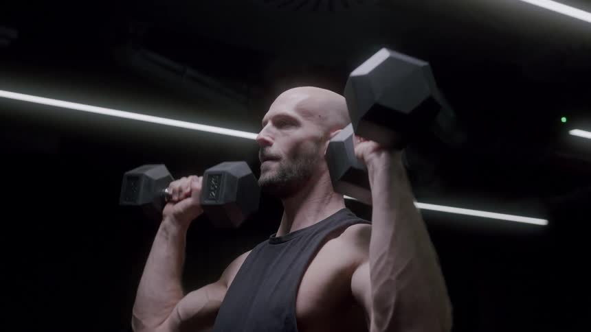 Man in a lowlight environment performing upward movements with dumbbells