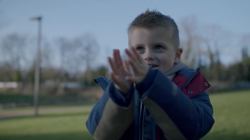 A toddler smiling and clapping while standing on a grass field during the day