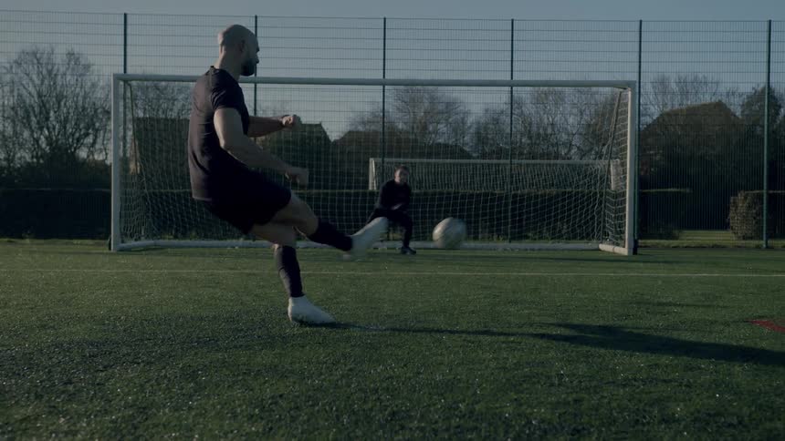 Man plays football kick towards the goal while the keeper dived to catch the ball which slipped under his arms