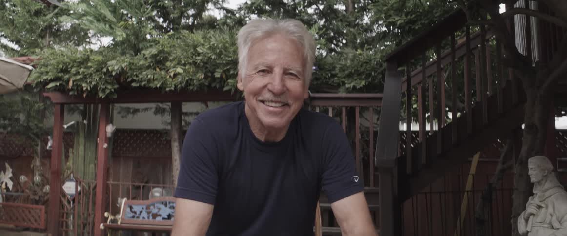 Elderly man wearing makeshift shoes while sitting in the backyard smiles at the camera