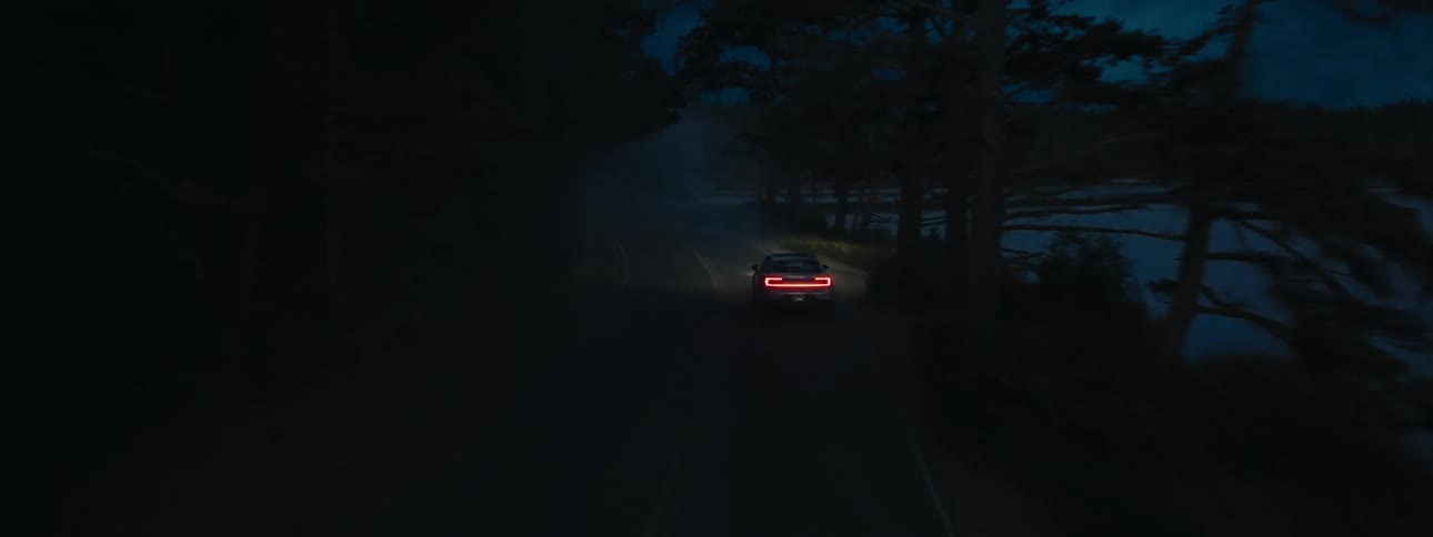 A car with its tail lights on is driving on a dark road beside a calm lake