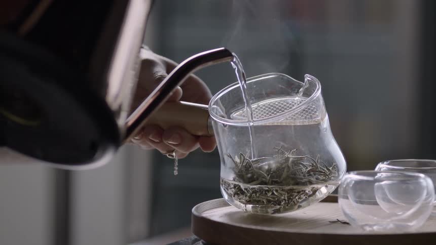 A person holding a glass mug and pouring water through a teapot that is falling onto a plant inside the jar