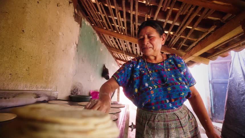 Older woman flips flat breads on large flat cooking surface under a wooden overhang