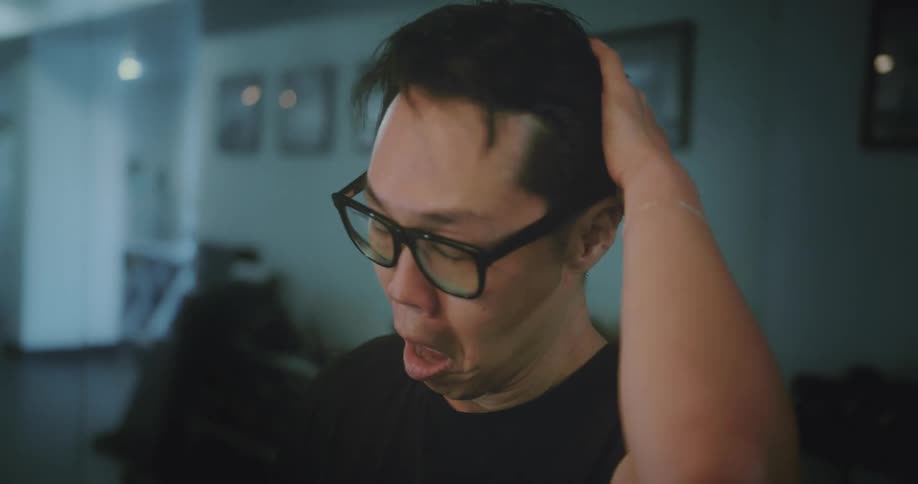Man standing in dimly lit room wipes his forehead and combs his hair back