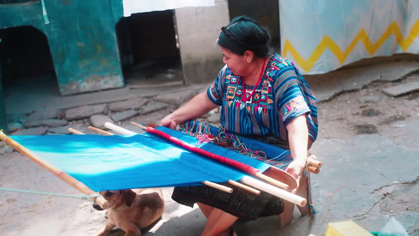 A woman is weaving a bright blue fabric with intricate designs on a loom