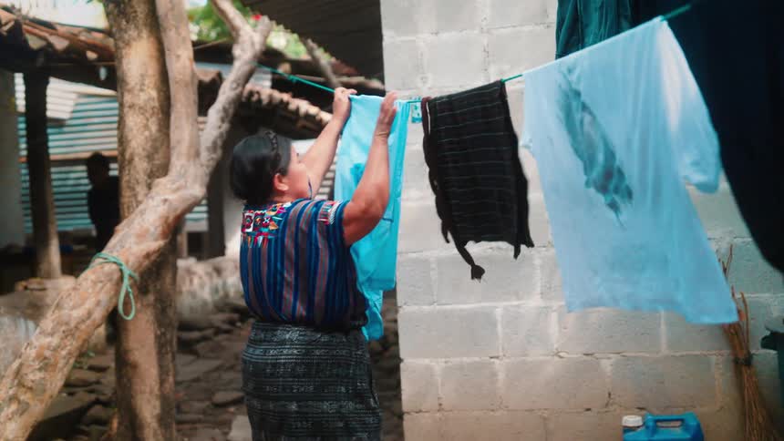 Older woman hangs clothes onto clothesline attached to tree between buildings