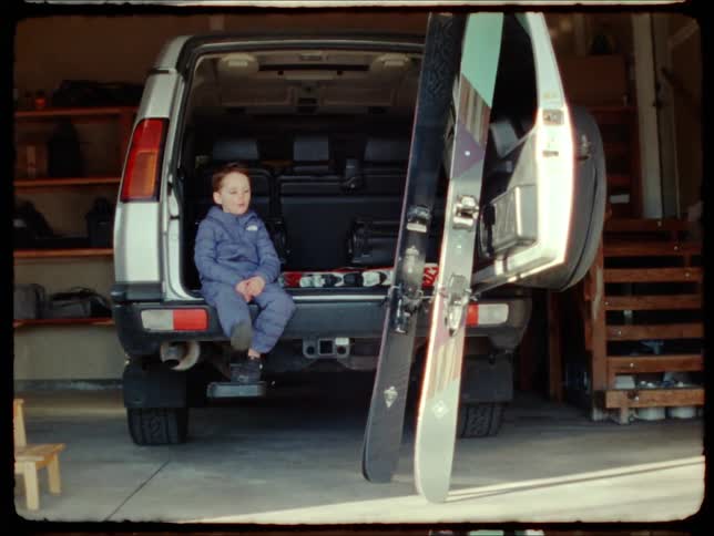 A boy in a blue outfit sits at the back of a car in the garage