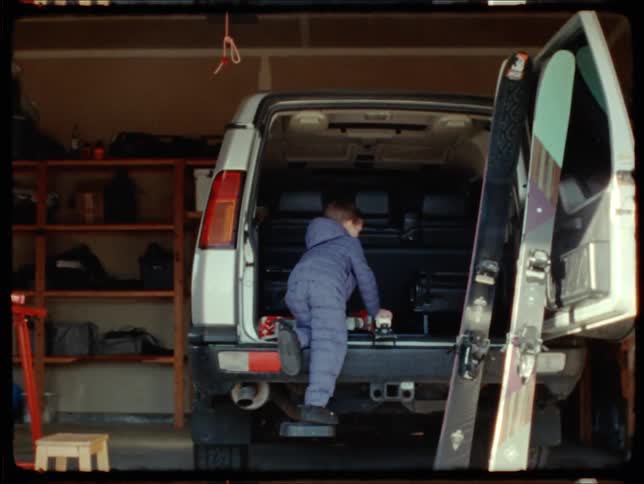 Young boy climbs into back of van parked in the garage with a pair of skis