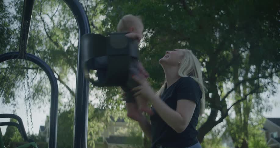 Mother pushes child in baby swing at playground in residential neighborhood