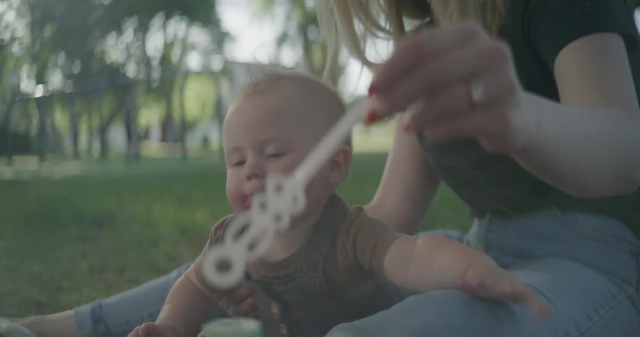 Mother sitting with baby on grass in park blows bubbles