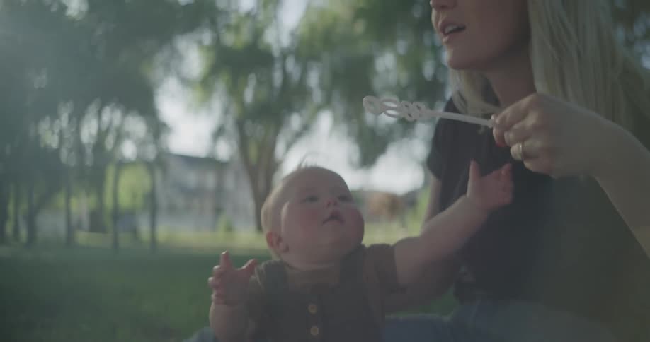 Mother sitting in a garden blowing a toy that blows soap bubbles and her baby son beside her