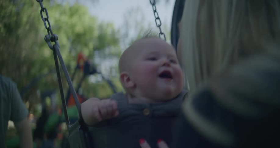 A man and a woman in a public park push their baby back and forth on a swing as the baby laughs