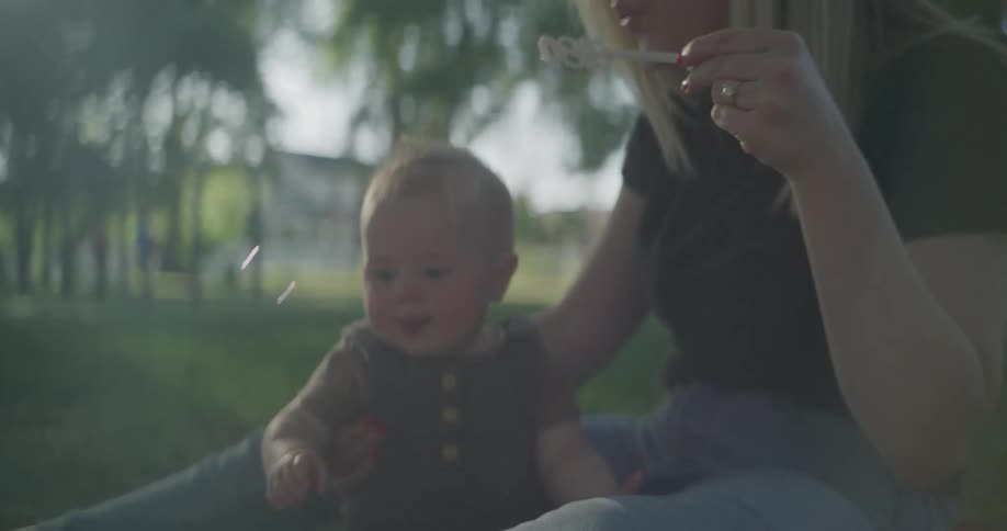 A woman sitting in the grass in a public park blows soap bubbles to entertain her baby