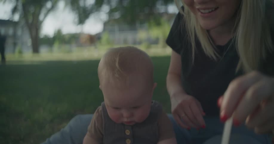 Young mother blows bubbles with a wand for her infant son as they sit on the grass under trees