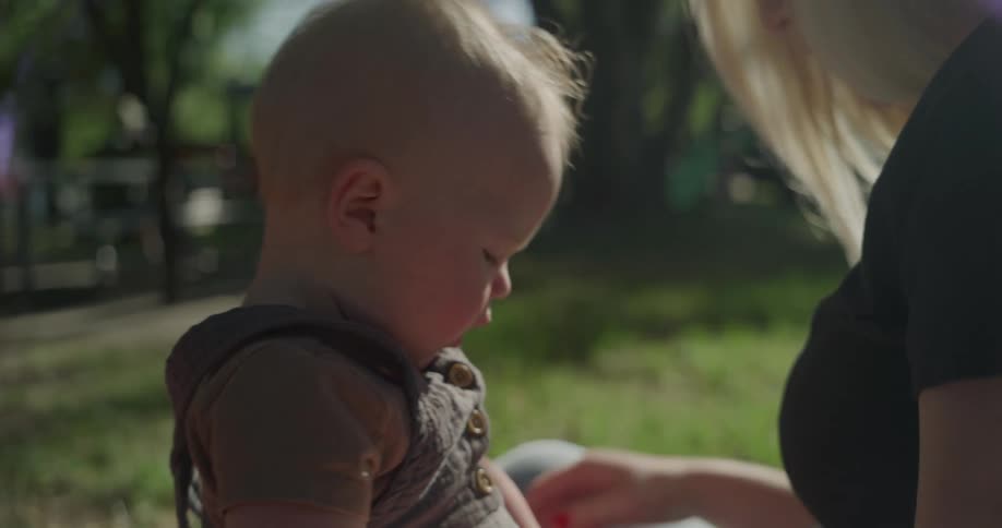 A mother and her infant son are sitting in the grass as she feeds him grapes