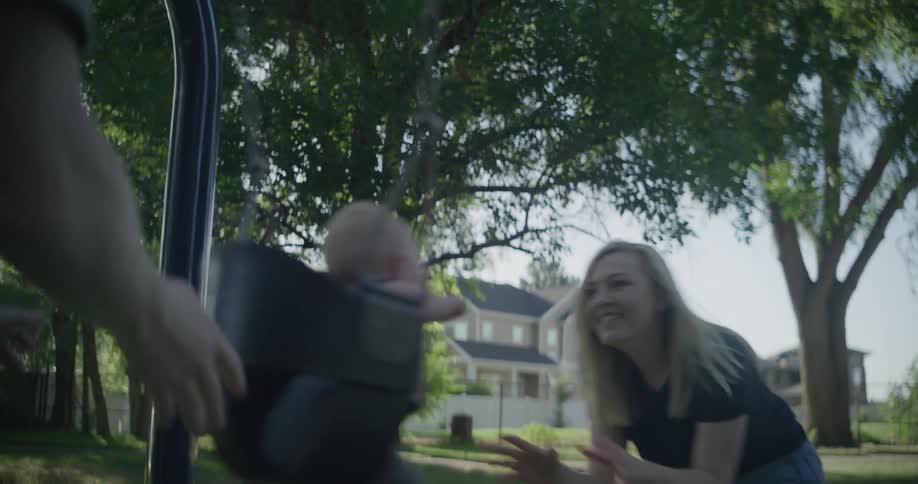 A man and a woman laughing and rocking a baby in a swing on a public park