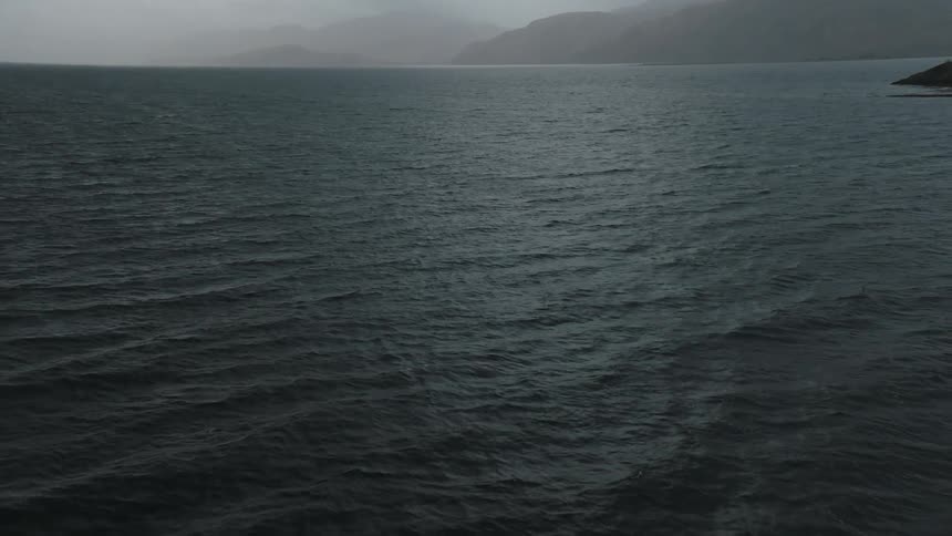 The water ripples and waves as dark storm clouds hang over mountains along the coast