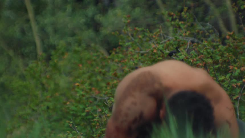 A naked man with spotted skin gets up from the ground in the woods