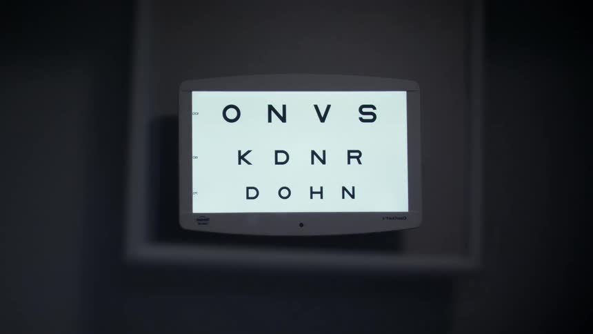 A digital screen shows an eye chart with letters of different sizes in a dark room