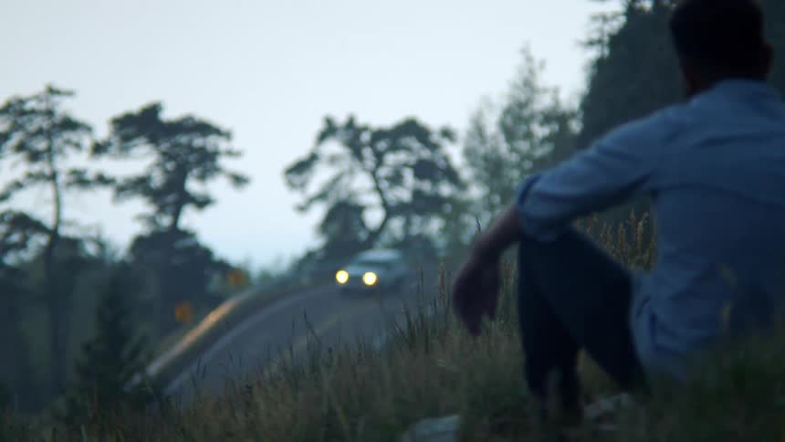 Man sitting on a hill facing a highway with a vehicle passing by