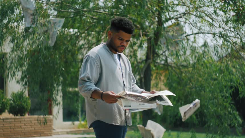 A man outdoors holds a newspaper with flying pages around him