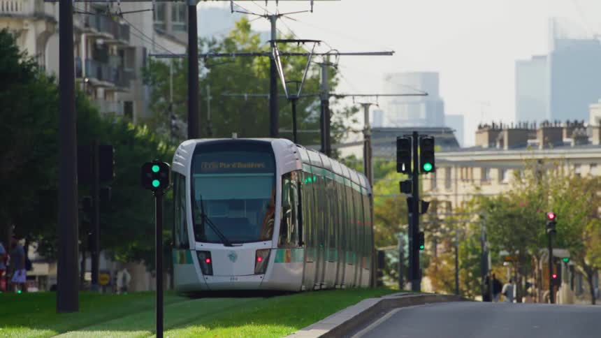 An electric train driving down the street on a city surrounded with trees