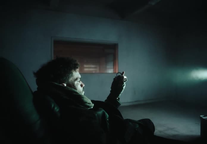Man sitting in a chair in an empty room holds object as bright light shines through window at night