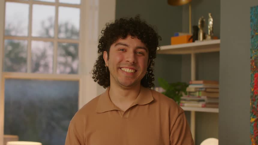 Man with curly hair earrings nose piercing and a brown polo shirt smiles at camera in a room