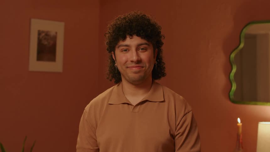 A man with curly hair looking forward and smiling and behind him there is an orange wall and a mirror