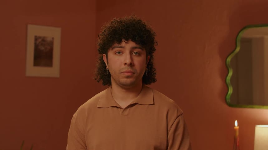 Man with curly hair and a brown polo shirt faces the camera with eye contact in the room with orange walls