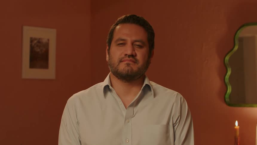 A man in a white shirt is standing looking forward and behind him there is an orange wall and a mirror
