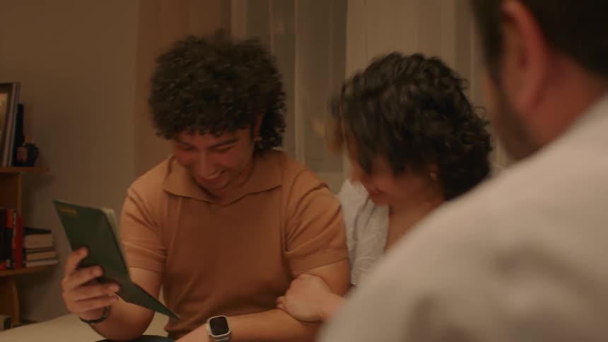 Two people sitting are showing a photo to a person in front of them and that person points to the photo and everyone laughs