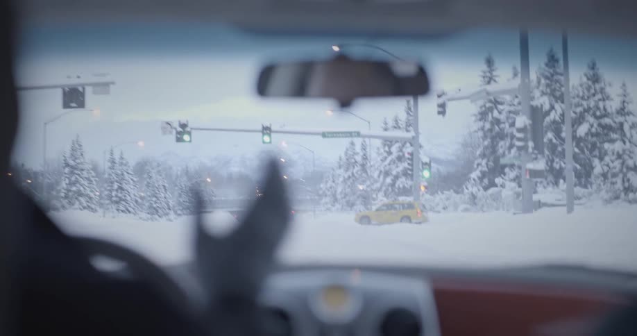 A backseat perspective through the front windshield of a vehicle approaching a green light at an intersection with everythingcovered in snow as the driver briefly takes one hand off the steering wheel