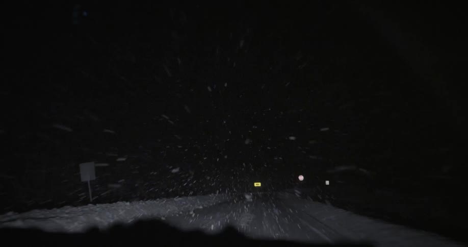 Late in the night the weather is snowing and a car drives on a lonely road with street sign seen on the side