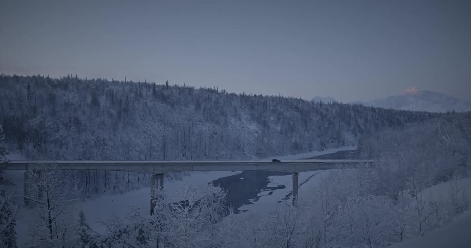 Vehicle crossing bridge over a frozen lake between two snow covered hills right before dark