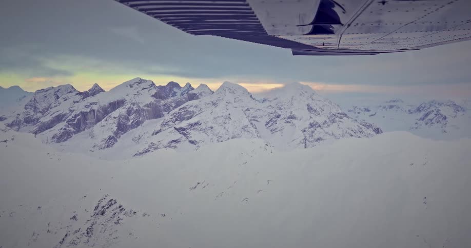 Airplane flies over snow covered mountains against a red tinted horizon