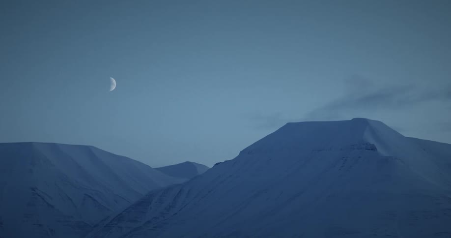 A half moon glows over snow covered mountains as clouds move across the sky